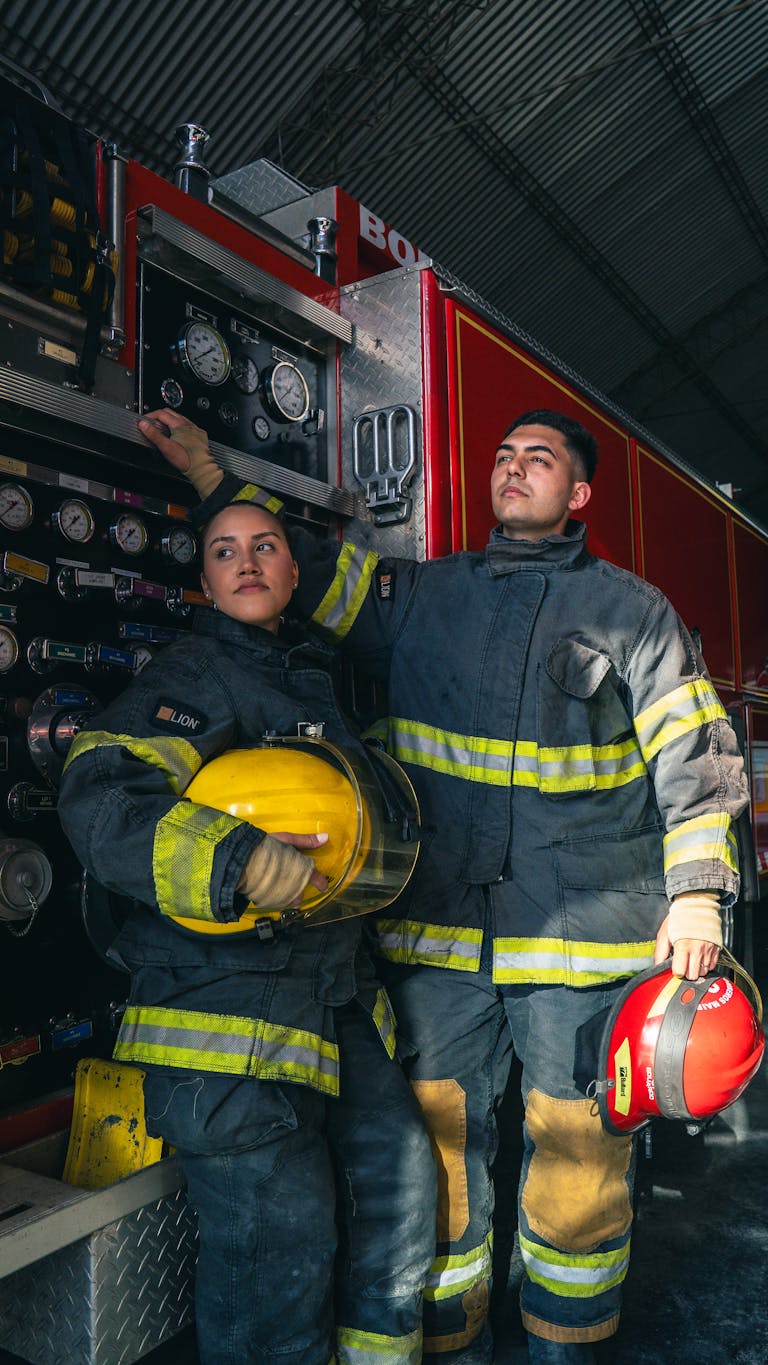 Two firefighters in uniform beside a firetruck, showing readiness and teamwork.